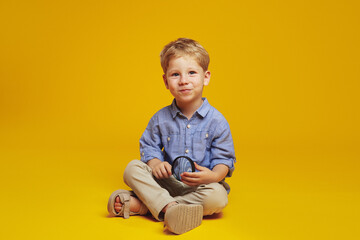 Little blonde haired boy in trendy blue shirt holding magnifier and looking at camera while sitting on studio floor with crossed legs, isolated over yellow background.