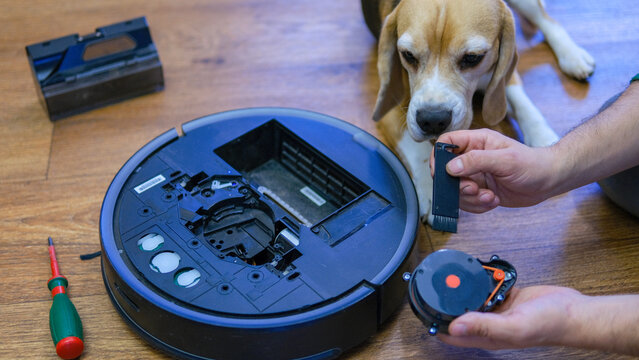 Caucasian young man with beagle dog doing vacuum cleaner robot maintenance. Male starts automatic vacuum cleaner. Robot vacuum cleaner repair.