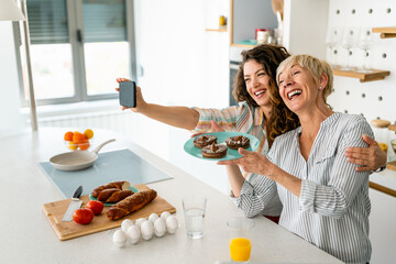 Two lovely smiling women taking selfie with mobile phone while holding plate of chocolate donuts in...