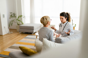 Senior blonde woman consulting with confident concentrated female doctor while sitting on sofa in...