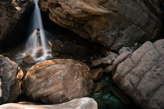 A young man in a blue t-shirt stands under a waterfall in a sandstone alcove