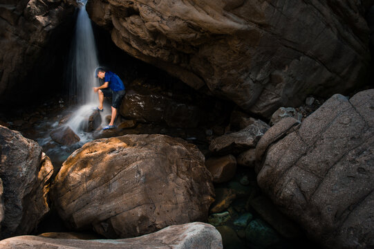 A young man in a blue t-shirt stands under a waterfall in a sandstone alcove