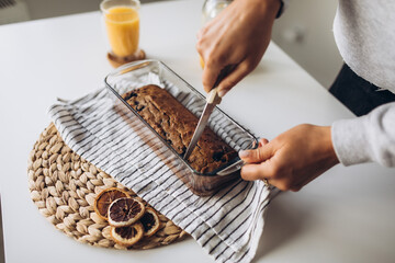 Close-up of a woman's hand cutting a freshly baked banana muffin with berries for breakfast along with sea buckthorn tea on the table - obrazy, fototapety, plakaty
