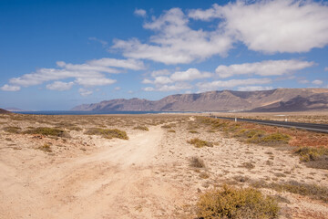 Desert landscape of white sand and desert bushes. Ocean and Famara cliff in the background. Dirt track. Sky with big white clouds. Lanzarote, Canary Islands, Spain.