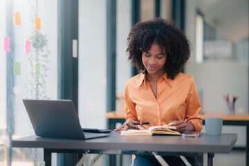 Young african american businesswoman using laptop computer, taking notes, working project in modern office. Financier working with documents at workplace.
