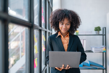 Young african american happy businesswoman standing and working on laptop by window in office.