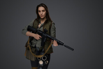 Fototapeta premium Middle Eastern-looking woman dressed in survivalist clothing posing with a rifle against a gray background, portraying strength and resilience