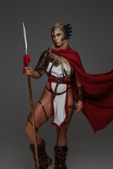 Viking woman adorned in fantasy armor, featuring a red cloak and exposed thighs. She confidently wields a spear on a gray backdrop