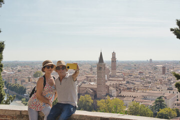 Happy married couple taking a selfie in front of the city of Verona,veneto. Two senior tourists having fun on a romantic summer vacation in Italy. Vacation concept and traveling lifestyle.