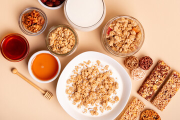 Healthy breakfast cereal. Granola with milk, glass of milk, honey, nuts and muesli bars with chocolate from above on light beige background.