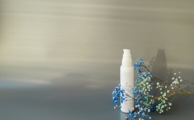 A white bottle with a cosmetic product on a gray background with diffused light and a sprig of blue flowers.  Space for copying text.  Concept of beauty sphere and beauty.