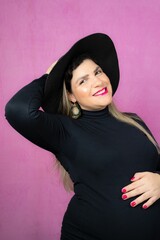 Pregnant Caucasian woman posing in a black dress for a maternity shoot on a pink background