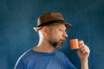 Man wearing a wide-brimmed hat and holding a bright orange mug filled posing in a studio