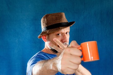 Man wearing a wide-brimmed hat and holding a bright orange mug filled posing in a studio