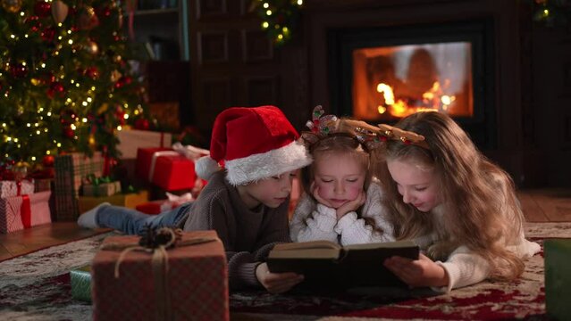 Merry Christmas Xmas holiday. Brother sisters children kids reading magic Christmas story book near Christmas tree in living room with fireplace in winter evening. Christmas eve at home