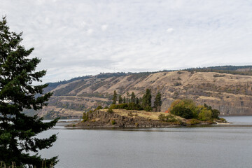 Fototapeta na wymiar Island on the Columbia river, Image shows the small island with a rock face and few trees on the calm river with a desert hill background