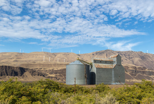 Grain silo and storage building by the Columbia river with the desert hills in the background where multiple wind turbines are spinning in the wind 