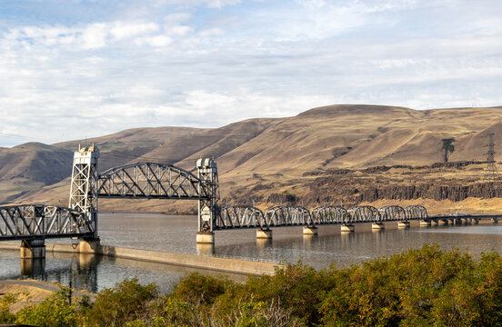 Bridge over the Columbia river, Image shows a bridge on a calm river reflecting the landscape off the water and steep hills with cloud and blue sky in the background