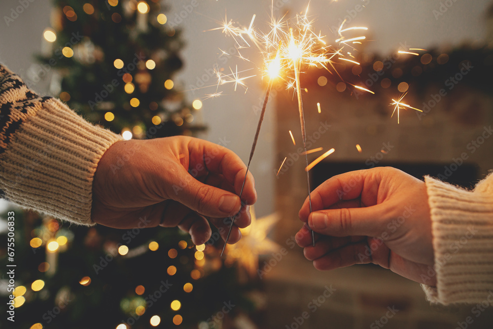 Wall mural Happy New Year! Burning sparklers in hands on background of modern country fireplace and christmas tree with golden lights. Fireworks glowing in hands, couple celebrating in festive decorated room