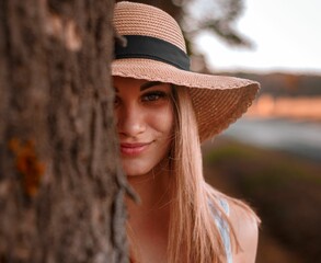 Young woman with long, golden blonde hair in  brown hat behind a tree