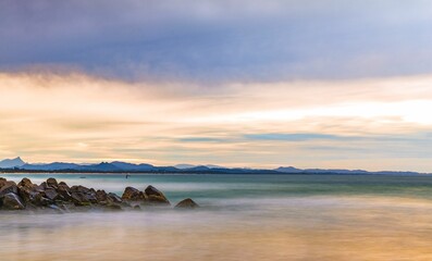 Long exposure shot of sandy beach shoreline against a backdrop of rocky outcrops and a cloudy sky.