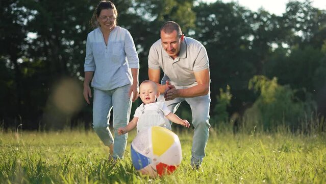 Happy family playing with ball on green meadow at sunset in the summer on vacation. Active family vacation picnic in nature on green grass. Parents teach the baby first steps by playing with the ball