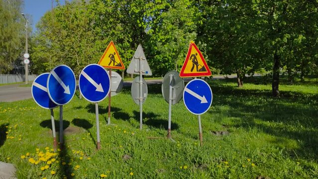 Many road signs stand together on grass in wrong place. Signs with arrows and road works are kept on green zone and confuse drivers on sunny summer day. Improper traffic management.