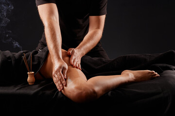 handsome male masseur doing a massage on a girl's leg on a black background, concept of therapeutic relaxing massage - obrazy, fototapety, plakaty