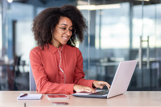 Young beautiful woman inside the office works with a laptop, a businesswoman in headphones listens music, podcasts, audio books and training course. Worker smiling , with curly hair and red shirt