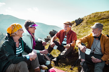 Group of senior friends sitting on mountain hill after long walk. Elderly hikers taking break from hike having drinks and snacks