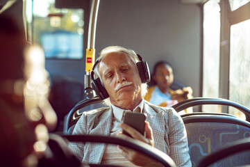 Senior man with headphones holding smartphone sitting on bus