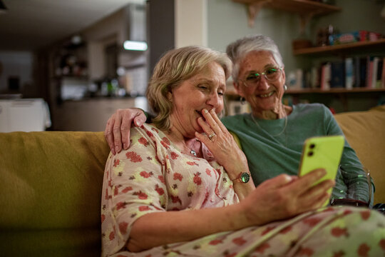 Elderly woman holding laugh with smartphone. Senior lesbian couple sitting on couch together at home