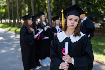 Group of happy students in graduation gowns outdoors. A young girl is holding a diploma.
