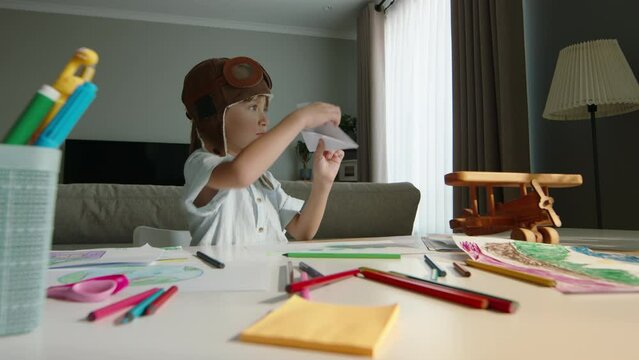 Concentrated little boy in pilot's cap makes airplane out of paper and launches it. Child dreams of becoming pilot and makes airplane while sitting at table. Fine motor skills development, creative