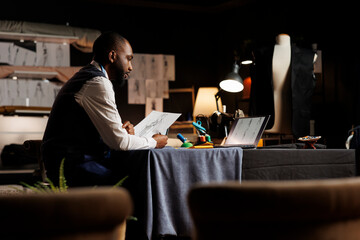 Hip level shot of skilled dressmaker in atelier shop workspace surrounded by sewing instruments looking online for design inspirations. Tailor studying drawings for upcoming fashion collection