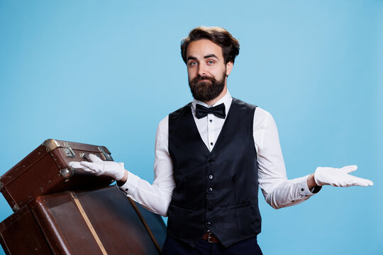Hotel concierge does i dont know gesture, shrugging in front of camera and acting uncertain next to trolley bags. Man bellhop feeling unsure and indicating doubt, unreliable answer.