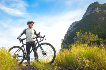 Obraz premium bearded cyclist stands with a bicycle against a background of blue sky and mountains. cycling and health-related hobbies
