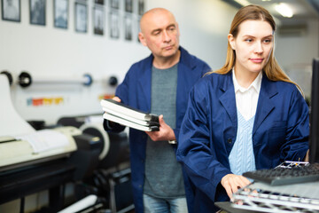 Woman in uniform, printing office worker, using computer, looking at display. Male co-worker standing beside her.