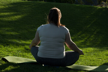 Portrait of overweight Caucasian middle-aged woman practicing yoga in nature. Fat woman meditating, relaxing in lotus position, sitting on fitness mat. Calm, relaxed, zen, stop stress. View from back