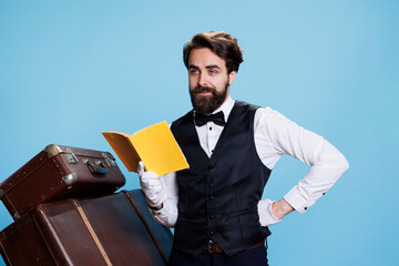 Hotel staff reads fiction book on camera, wearing tie and gloves while he is enjoying literature against blue background. Employee having fun with lecture hobby in studio, novel story.