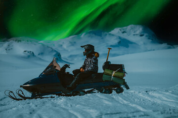Scenic close up side photo of girl sitting on snow mobile with night mountains and northern green...