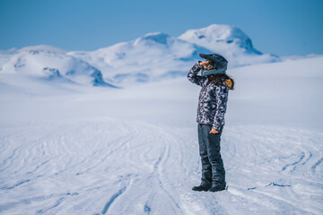 Girl wearing snowmobile helmet, sun glasses and warm overall looking forward with hand over eyes,...