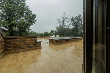 Terrace of a residential home with a white coffee table and matching metal chairs, trees and a lot of rain falling from the sky