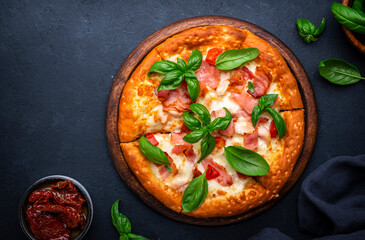 Homemade hot pizza with smoked ham, cheese, spicy tomato sauce and green basil on black table background, top view