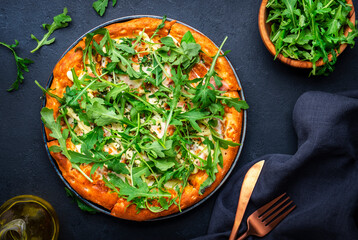 Hot homemade pizza with blue cheese, ham, pesto sauce and fresh arugula, black table background, top view