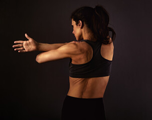 Female sporty muscular with ponytail doing stretching workout of the shoulders, blades in sport bra, holding the hands on the shoulders on black background with empty copy space.