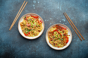 Two bowls with Chow Mein or Lo Mein, traditional Chinese stir fry noodles with meat and vegetables, served with chopsticks top view on rustic blue concrete background. - 675581689