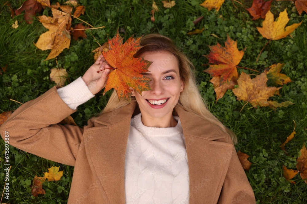 Sticker Smiling woman lying on grass and covering eye with autumn leaf, top view