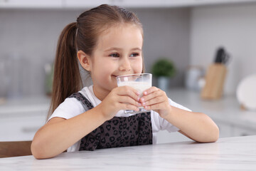 Cute girl drinking fresh milk from glass at white table in kitchen