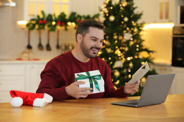 Celebrating Christmas online with exchanged by mail presents. Happy man with greeting card and gift box during video call on laptop at home